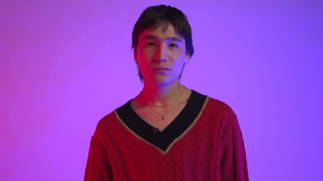 Miruh trägt einen roten V-Ausschnittpulli mit schwarzem Kragen und eine feine Kette, hat dunkle Haare mit Mittelscheitel. Miruh trägt einen roten V-Ausschnittpulli mit schwarzem Kragen und eine feine Kette, hat dunkle Haare mit Mittelscheitel.
