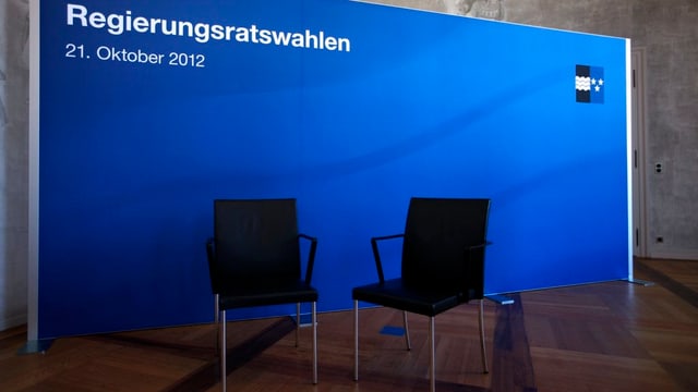 Zwei leere Stühle vor einer Leinwand mit dem Aufdruck "Regierungsratswahlen" und dem Aargauer Wappen (Symbolbild). Zwei leere Stühle vor einer Leinwand mit dem Aufdruck "Regierungsratswahlen" und dem Aargauer Wappen (Symbolbild).