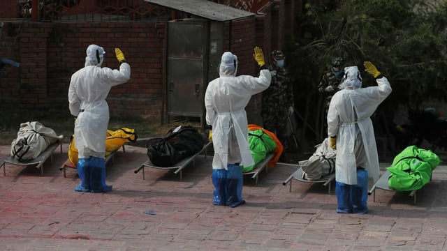 Männer in Schutzanzügen salutieren vor den aufgebahrten, in bunte Tücher gewickelte Leichen. Männer in Schutzanzügen salutieren vor den aufgebahrten, in bunte Tücher gewickelte Leichen.