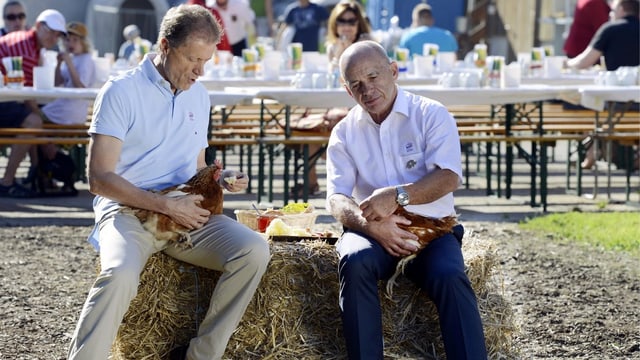 Migros-CEO Bolliger und Bundesrat Maurer sitzen auf einem Strohballen und halten je ein Huhn fest.