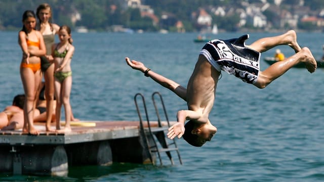 Ein Junge springt in den See, per Salto, auf einem Schwimmfloss schauen Mädchen ihm zu. Ein Junge springt in den See, per Salto, auf einem Schwimmfloss schauen Mädchen ihm zu.
