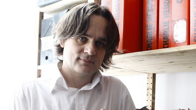 Porträt eines Mannes vor einem Regal mit Charlie Hebdo Sammlungen. Porträt eines Mannes vor einem Regal mit Charlie Hebdo Sammlungen.