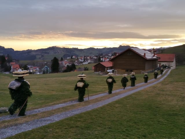 Auf einem Feldweg marschieren verkleidete Gestalten. Im Hintergrund Berge mit Aufhellung.