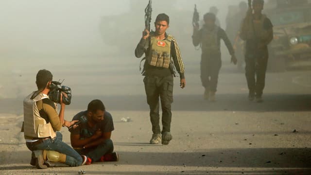 Ein Journalist kniet mit der Kamera neben einem verletzten Mann. Mehrere schwer bewaffnete Männer gehen an ihnen vorüber. Ein Journalist kniet mit der Kamera neben einem verletzten Mann. Mehrere schwer bewaffnete Männer gehen an ihnen vorüber.