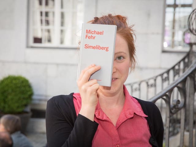 Eine junge Frau hält sich ein graues Buch vor das Gesicht.