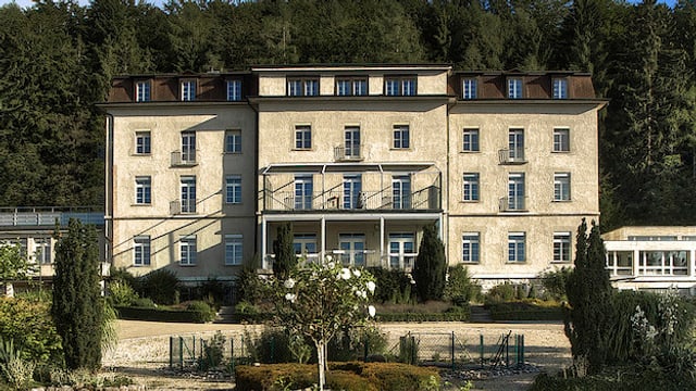 Bild von der Fridau von aussen, Frontalaufnahme. Es ist ein grosses, altes, längliches, beiges Gebäude, das links und rechts Nebentrakte hat. Im Hintergrund sieht man einen Wald. Bild von der Fridau von aussen, Frontalaufnahme. Es ist ein grosses, altes, längliches, beiges Gebäude, das links und rechts Nebentrakte hat. Im Hintergrund sieht man einen Wald.