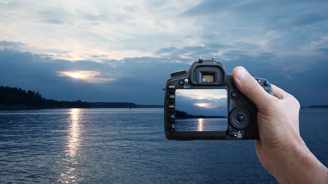 Ein Mann fotografiert einen Sonnenuntergang am See und betrachtet die Aufnahme im Kamera-Display