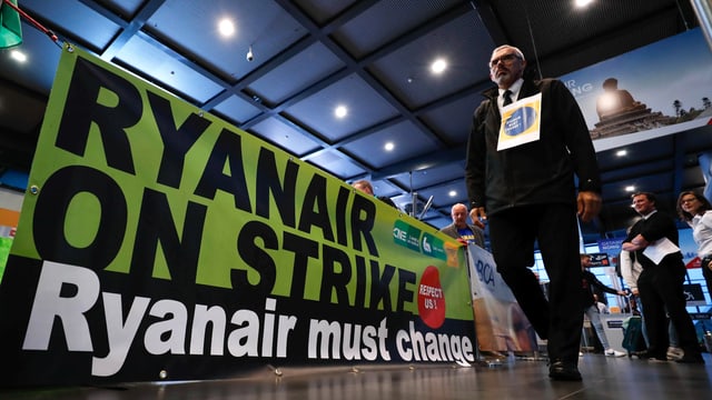 Pilot mit Schild um den Hals läuft an einem Plakat vorbei. Darauf steht: Ryanair on Strike. Ryanair must change. Pilot mit Schild um den Hals läuft an einem Plakat vorbei. Darauf steht: Ryanair on Strike. Ryanair must change.