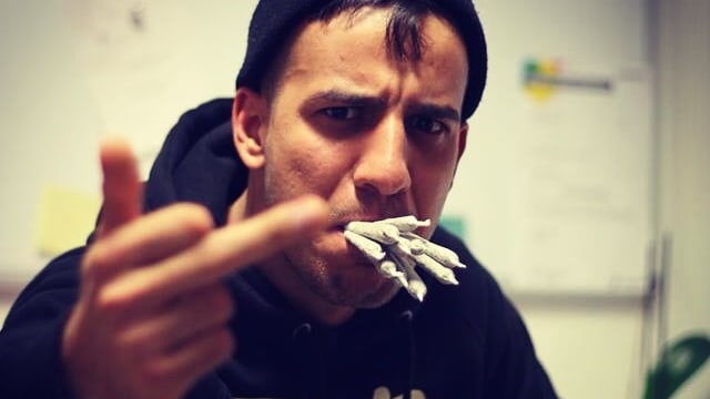 Böse guckender Rapper ist zu sehen, mit gerecktem Mittelfinger und einem Mund voll Joints