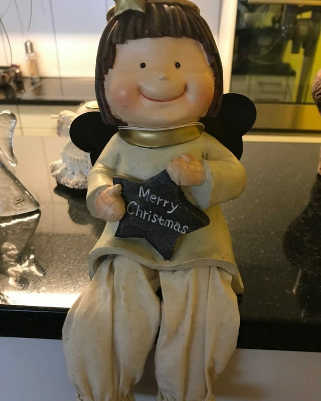 Grinsender Porzellanengel mit schwarzen Flügeln und einem Stern in der Hand auf dem Merry Christmas steht.