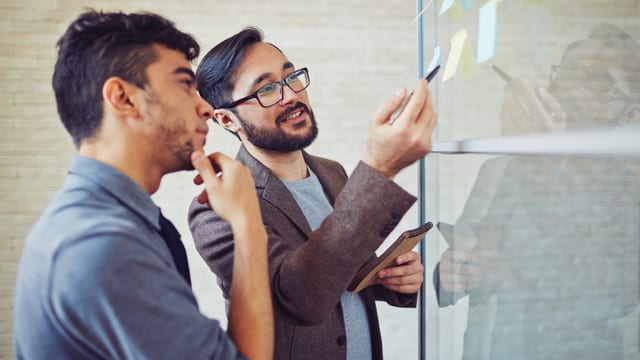 Zwei männer besprechen die Notizen auf einem Post-it. Der eine lächelt und erklärt, während der andere zuhört. Zwei männer besprechen die Notizen auf einem Post-it. Der eine lächelt und erklärt, während der andere zuhört.