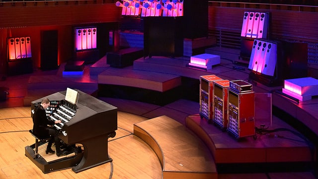 Ein Mann sitzt an einer Orgel, im Hintergrund grellpinke Lautsprecher. Ein Mann sitzt an einer Orgel, im Hintergrund grellpinke Lautsprecher.