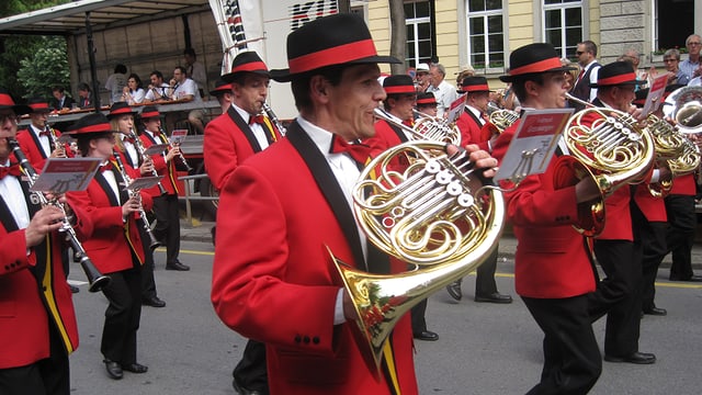Musicants en giaccas cotschnas e cun chapella, che sunan durant marschar atras ina via da la citad da Cuira. Musicants en giaccas cotschnas e cun chapella, che sunan durant marschar atras ina via da la citad da Cuira.