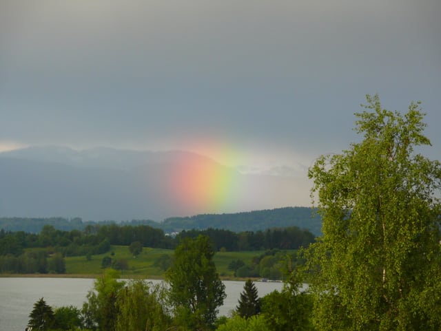 See mit Regenbogen. In der Umgebung grüne Bäume und Wiesen. Im Hintergrund Berge mit Schnee.