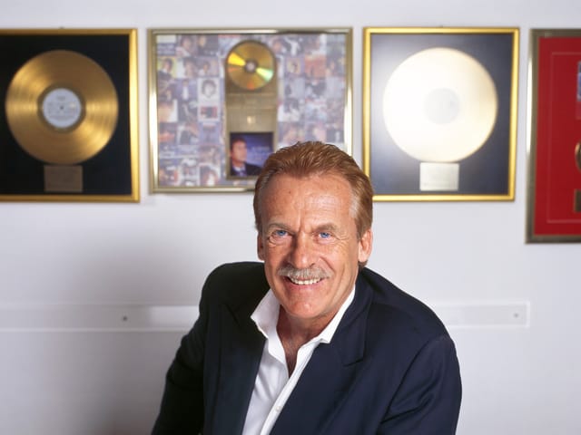 Ein Mann im weissen Hemd und dunklen Jackett sitzt vor einer Wand, an der goldene Schallplatten hängen. Ein Mann im weissen Hemd und dunklen Jackett sitzt vor einer Wand, an der goldene Schallplatten hängen.