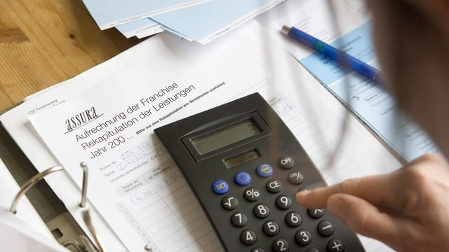 Junge Frau arbeitet mit Taschenrechner, dahinter Ordner mit einem Schreiben der Assura Junge Frau arbeitet mit Taschenrechner, dahinter Ordner mit einem Schreiben der Assura