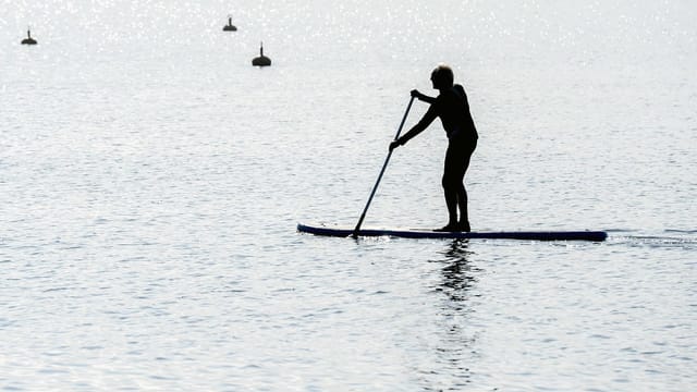 Ein Mann steht auf einem Brett, einem Stand-Up-Paddle auf dem Pfäffikersee. In der Hand hält er ein Paddel. Ein Mann steht auf einem Brett, einem Stand-Up-Paddle auf dem Pfäffikersee. In der Hand hält er ein Paddel.