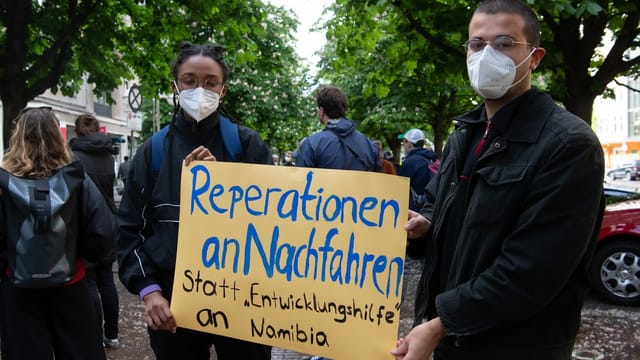 Junge Fraue und junger Mann mit Schild. Aufschrift: Reparation statt Entwicklungshilfe Junge Fraue und junger Mann mit Schild. Aufschrift: Reparation statt Entwicklungshilfe
