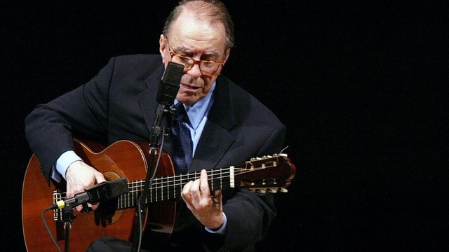 João Gilberto hät eine Gitarre in der Hand und singt ins Mikrofon, das vor ihm steht. João Gilberto hät eine Gitarre in der Hand und singt ins Mikrofon, das vor ihm steht.