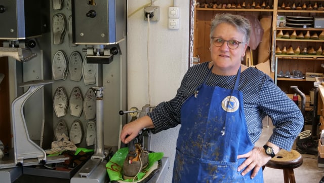Eine Frau mit kurzen grauen Haaren und Brille. Sie trägt eine blaue Schürze und steht in einer Schumacherei. Eine Frau mit kurzen grauen Haaren und Brille. Sie trägt eine blaue Schürze und steht in einer Schumacherei.