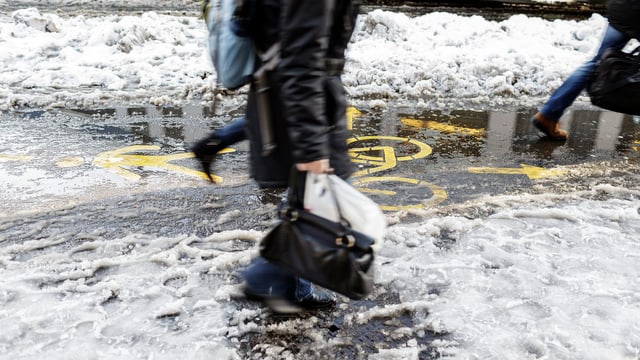 Eine Frau fährt mit dem Fahrrad über eine Strasse. Es schneit. Im Vodergrund ein Baum mit Schnee. Eine Frau fährt mit dem Fahrrad über eine Strasse. Es schneit. Im Vodergrund ein Baum mit Schnee.