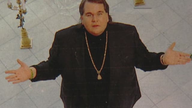 Ein übergewichtiger Mann mit goldenem Amulett steht mit ausgebreiteten Armen da und schaut nach oben in die Kamera. Ein übergewichtiger Mann mit goldenem Amulett steht mit ausgebreiteten Armen da und schaut nach oben in die Kamera.