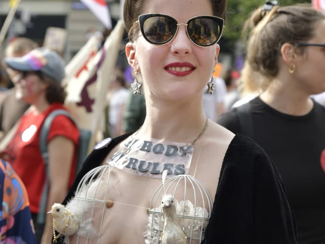 Eien Frau trägt zwei Vogelkäfige auf ihren Brüsten und ein Schild "My Body My Rules" am Hals. Eien Frau trägt zwei Vogelkäfige auf ihren Brüsten und ein Schild "My Body My Rules" am Hals.