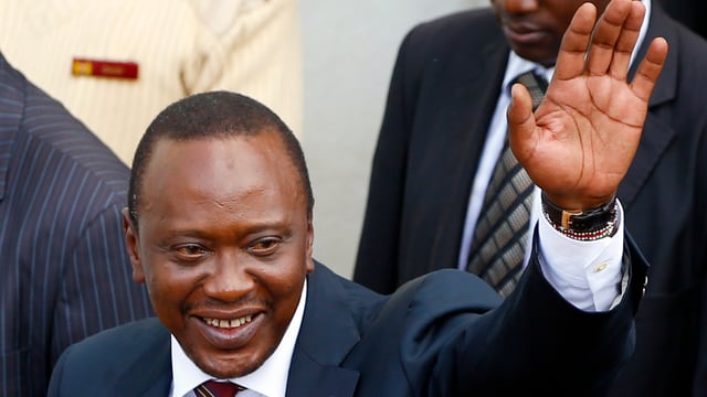 Kenyatta, im Anzug, hält eine Hand zu Gruss hoch. Kenyatta, im Anzug, hält eine Hand zu Gruss hoch.