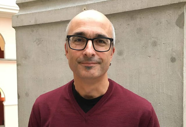 Ein Mann mit Brille steht vor einer Säule und lächelt mit geschlossenem Mund in die Kamera. Ein Mann mit Brille steht vor einer Säule und lächelt mit geschlossenem Mund in die Kamera.
