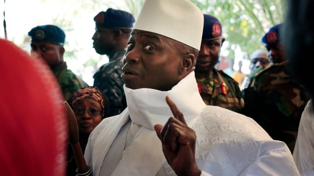 Jammeh in Weiss blickt in die Kamera mit erhobenem Zeigfinger. Jammeh in Weiss blickt in die Kamera mit erhobenem Zeigfinger.