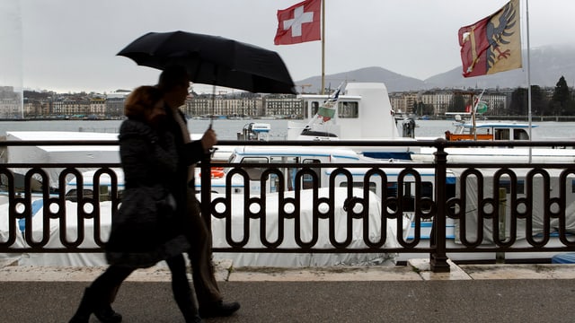 Ein Paar geht am Genferseeufer entlang. Er hält einen Regenschirm. Auf Booten wehen Schweiz- und Genffahnen. Ein Paar geht am Genferseeufer entlang. Er hält einen Regenschirm. Auf Booten wehen Schweiz- und Genffahnen.