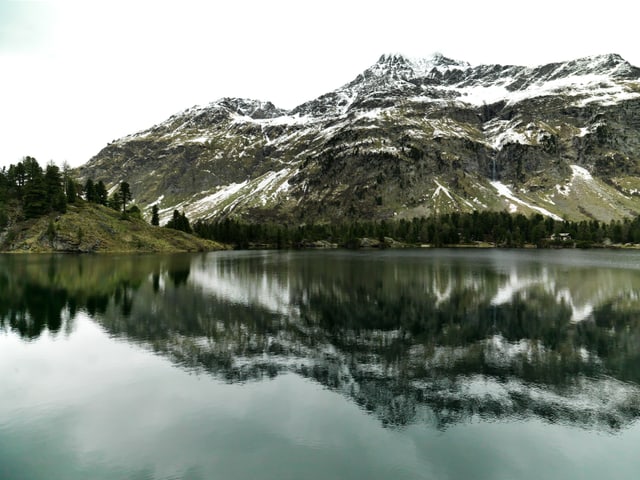 Berge spiegeln sich im See, darüber bewölkter Himmel.