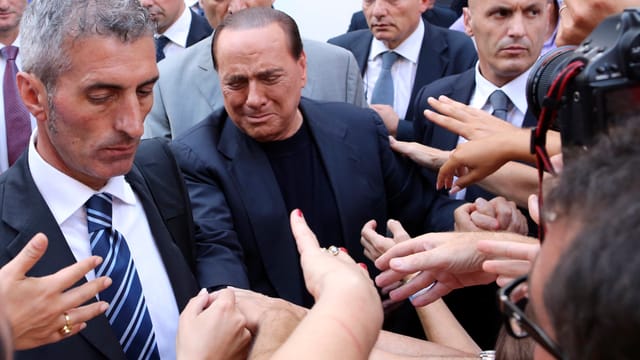 Ein zerknirschter Berlusconi inmitten von Bodyguards und Menschen, die ihm die Hand schütteln wollen. Ein zerknirschter Berlusconi inmitten von Bodyguards und Menschen, die ihm die Hand schütteln wollen.