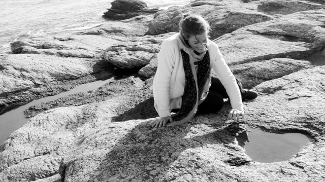Eine Frau sitzt am Meer auf einem flachen Felsen, durch den Stein haben sich kleine Bäche gefressen. Die Frau sitzt vor einer Wasserpfütze und lächelt. Sie trägt die Haare locker zusammengebunden, einen Schal und eine Strickjacke. Eine Frau sitzt am Meer auf einem flachen Felsen, durch den Stein haben sich kleine Bäche gefressen. Die Frau sitzt vor einer Wasserpfütze und lächelt. Sie trägt die Haare locker zusammengebunden, einen Schal und eine Strickjacke.
