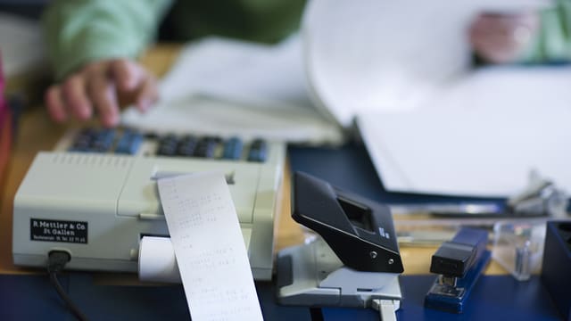 Eine Person benutzt einen Tischrechner und blättert zugleich in Unterlagen. Eine Person benutzt einen Tischrechner und blättert zugleich in Unterlagen.