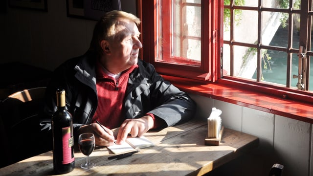 Hanns-Josef Ortheil sitzt mit einem Glas Wein und Notitzbuch an einem Tisch und schaut aus dem Fenster. Hanns-Josef Ortheil sitzt mit einem Glas Wein und Notitzbuch an einem Tisch und schaut aus dem Fenster.
