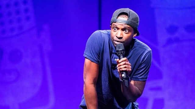 Charles Nguela steht bei der Comedy Talent Show auf der Bühne, hat ein Mikrofon in der Hand und schaut ausdrucksvoll in das Publikum. Charles Nguela steht bei der Comedy Talent Show auf der Bühne, hat ein Mikrofon in der Hand und schaut ausdrucksvoll in das Publikum.