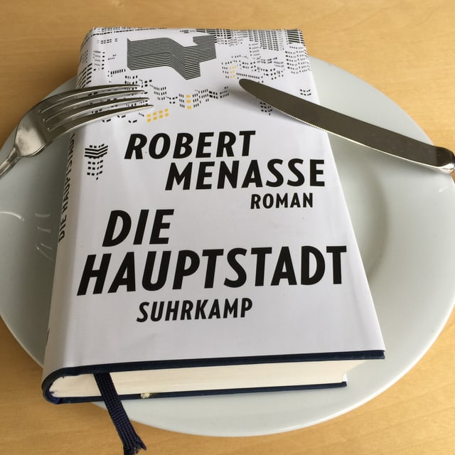 Der Roman «Die Hauptstadt» von Robert Menasse liegt auf einem weissen Teller. Messer und Gabel angelehnt. Der Roman «Die Hauptstadt» von Robert Menasse liegt auf einem weissen Teller. Messer und Gabel angelehnt.
