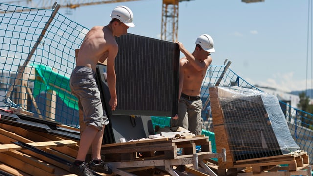 Zwei junge Männer heben auf einem Dach ein Solarpanel vom Palett Zwei junge Männer heben auf einem Dach ein Solarpanel vom Palett