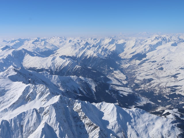 Hoch über den verschneiten Berggipfel und Täler herrscht brilliante Fernsicht. Der Himmel ist blau.