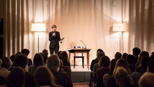 Eine Frau steht mit dem Mikrofon auf der Bühne, vor ihr ist ein Publikum zu sehen. Eine Frau steht mit dem Mikrofon auf der Bühne, vor ihr ist ein Publikum zu sehen.