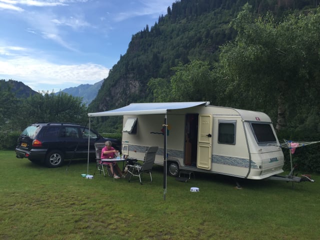 Wohnwagen mit aufgeklapptem Vordach, darunter sitzt eine Frau. Weiter hinten steht ein Auto mit holländischem Kontrollschild. Tessiner Berglandschaft.