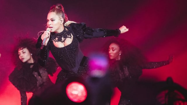 Eine Frau in schwarzem Kleid steht singend mit Mikrofon auf einer Bühne, die in dunkelrotes Licht getaucht ist. Eine Frau in schwarzem Kleid steht singend mit Mikrofon auf einer Bühne, die in dunkelrotes Licht getaucht ist.