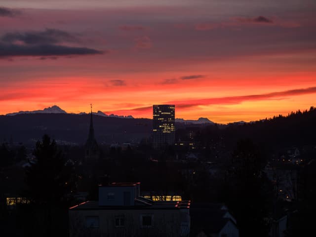 Im Vordergrund sieht man dunkle Gebäude und ein Hochhaus mit erhellten Fenstern. Im Hintergrund ist der Himmel orange bis rot gefärbt.