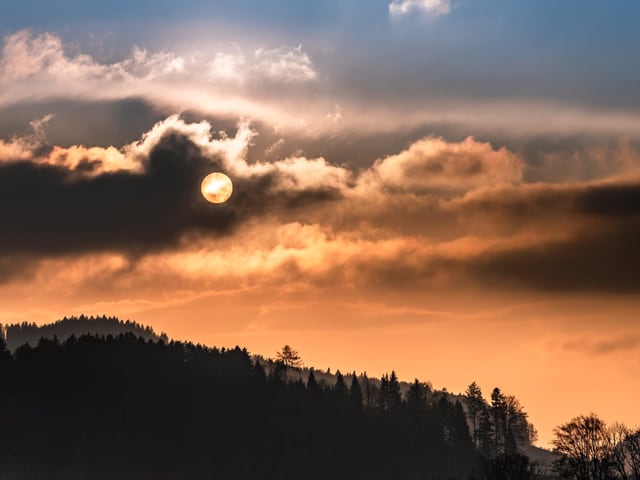 Bewölkter Himmel in gelb bis grau-blau. Hinter einer Wolke ist die Sonnenscheibe zu erkennen.