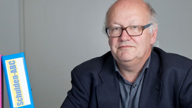 Mario Roncoroni sitzt an einem Tisch, daneben steht ein Ordner mit der Aufschrift «Schulden-ABC» Mario Roncoroni sitzt an einem Tisch, daneben steht ein Ordner mit der Aufschrift «Schulden-ABC»