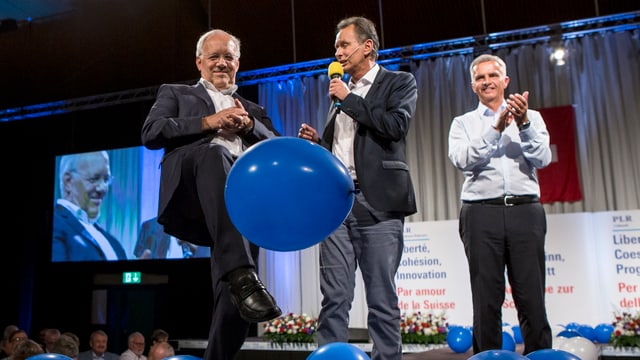 Johann Schneider-Ammann, Philipp Müller und Didier Burkhalter auf der Bühne. Schneider-Ammann kickt einen blauen Ballon weg. Johann Schneider-Ammann, Philipp Müller und Didier Burkhalter auf der Bühne. Schneider-Ammann kickt einen blauen Ballon weg.