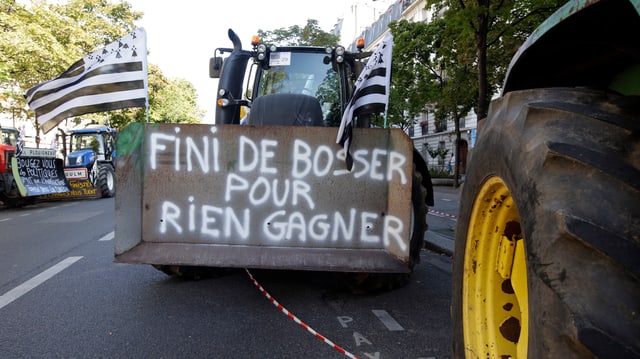 Ein Traktor mit schwarz-weiss gestreiften Fahnen und einem Schild vorne: Fini de bosser pour rien gagner. Ein Traktor mit schwarz-weiss gestreiften Fahnen und einem Schild vorne: Fini de bosser pour rien gagner.