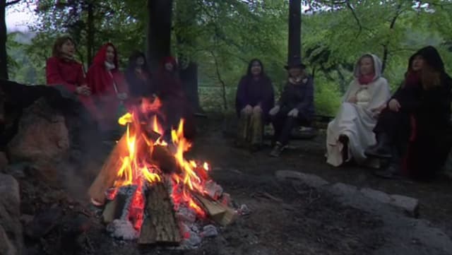 Feuer im Wald, dahinter sitzt eine Runde von Frauen in farbigen Umhängen Feuer im Wald, dahinter sitzt eine Runde von Frauen in farbigen Umhängen