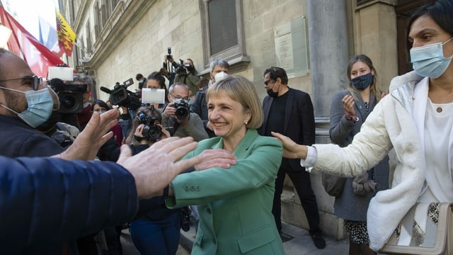 Frau mit grüner Jacke begrüsst jubelnde Anhänger. Medienleute mit Kamera filmen. Dahinter Genfer Fahne. Frau mit grüner Jacke begrüsst jubelnde Anhänger. Medienleute mit Kamera filmen. Dahinter Genfer Fahne.